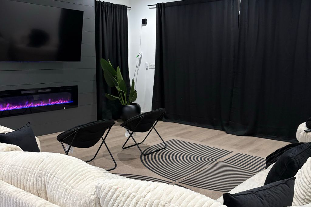 A modern living room with a flat-screen TV, a fireplace, black curtains, a large potted plant, and a patterned rug.