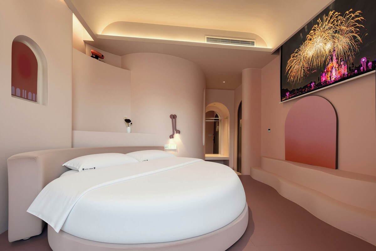 Un dormitorio moderno con una cama redonda, una lámpara fijada a la pared y un ventanal con vistas a fuegos artificiales.