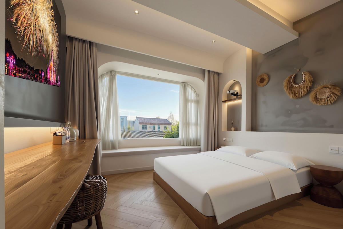 Una habitación de hotel moderna con una cama grande, un escritorio de madera, una ventana con cortinas y un televisor montado en la pared mostrando fuegos artificiales.