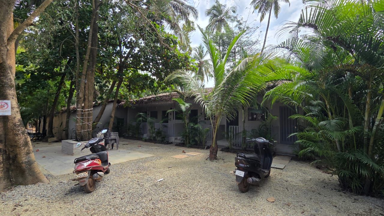 A courtyard with two scooters parked, surrounded by trees and a building with a thatched roof.