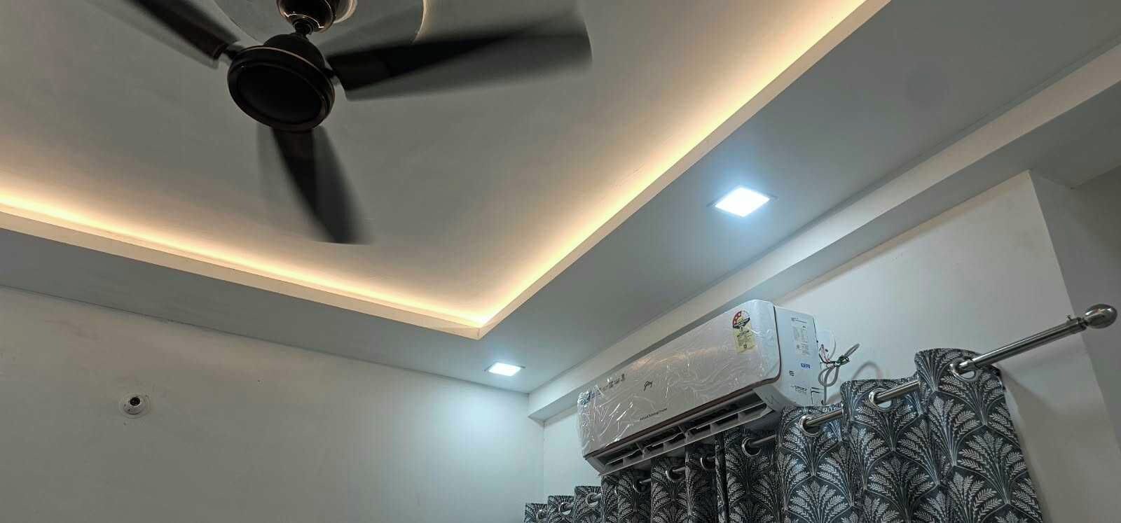 A ceiling fan, recessed lighting, and an air conditioning unit in a room with patterned curtains.
