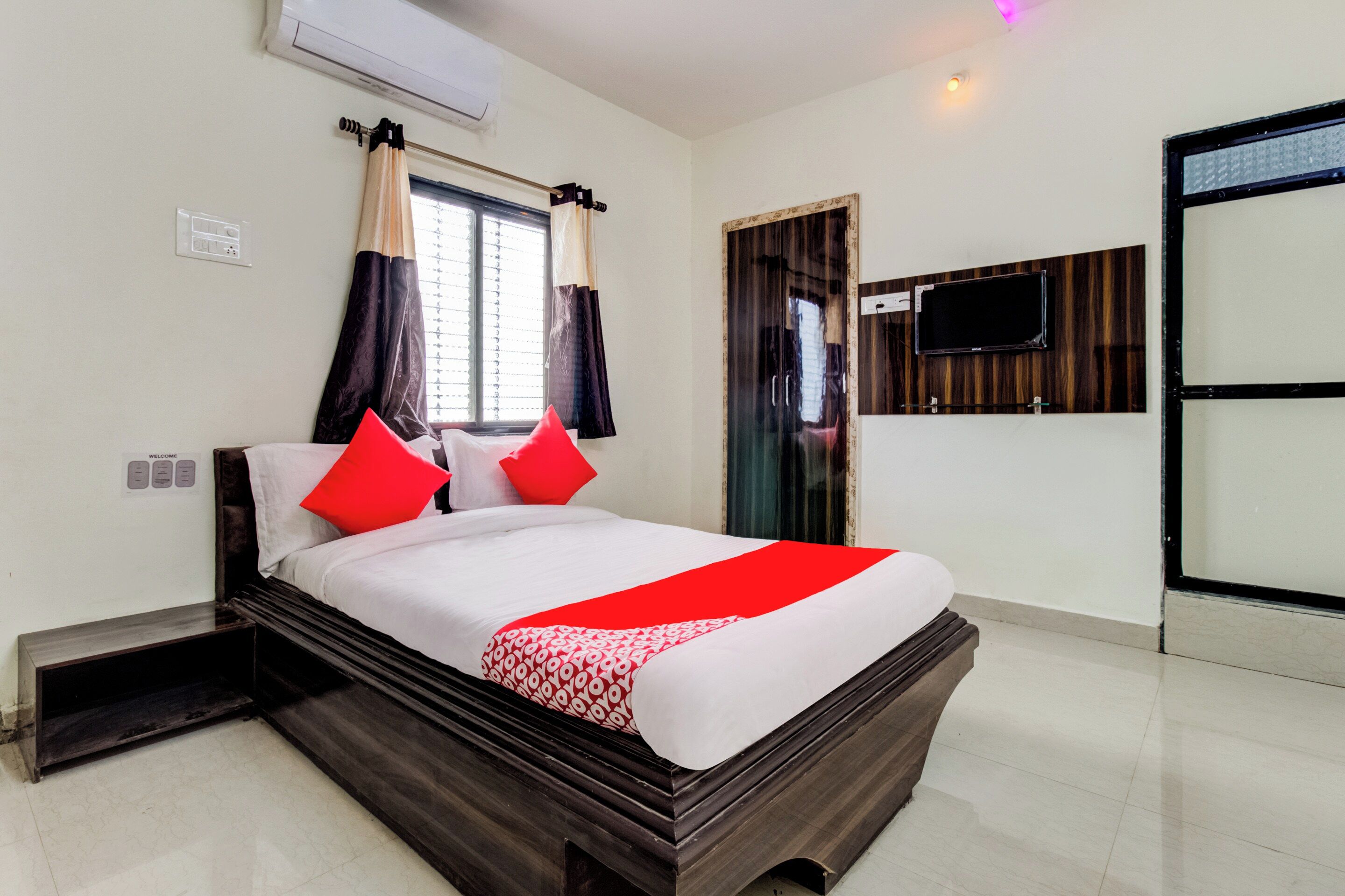 A hotel room with a large bed, a flat-screen TV mounted on the wall, and a window with curtains.