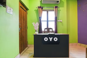 A reception desk with 'OYO' branding, a wooden door, a window with curtains, and a wall-mounted air conditioner.