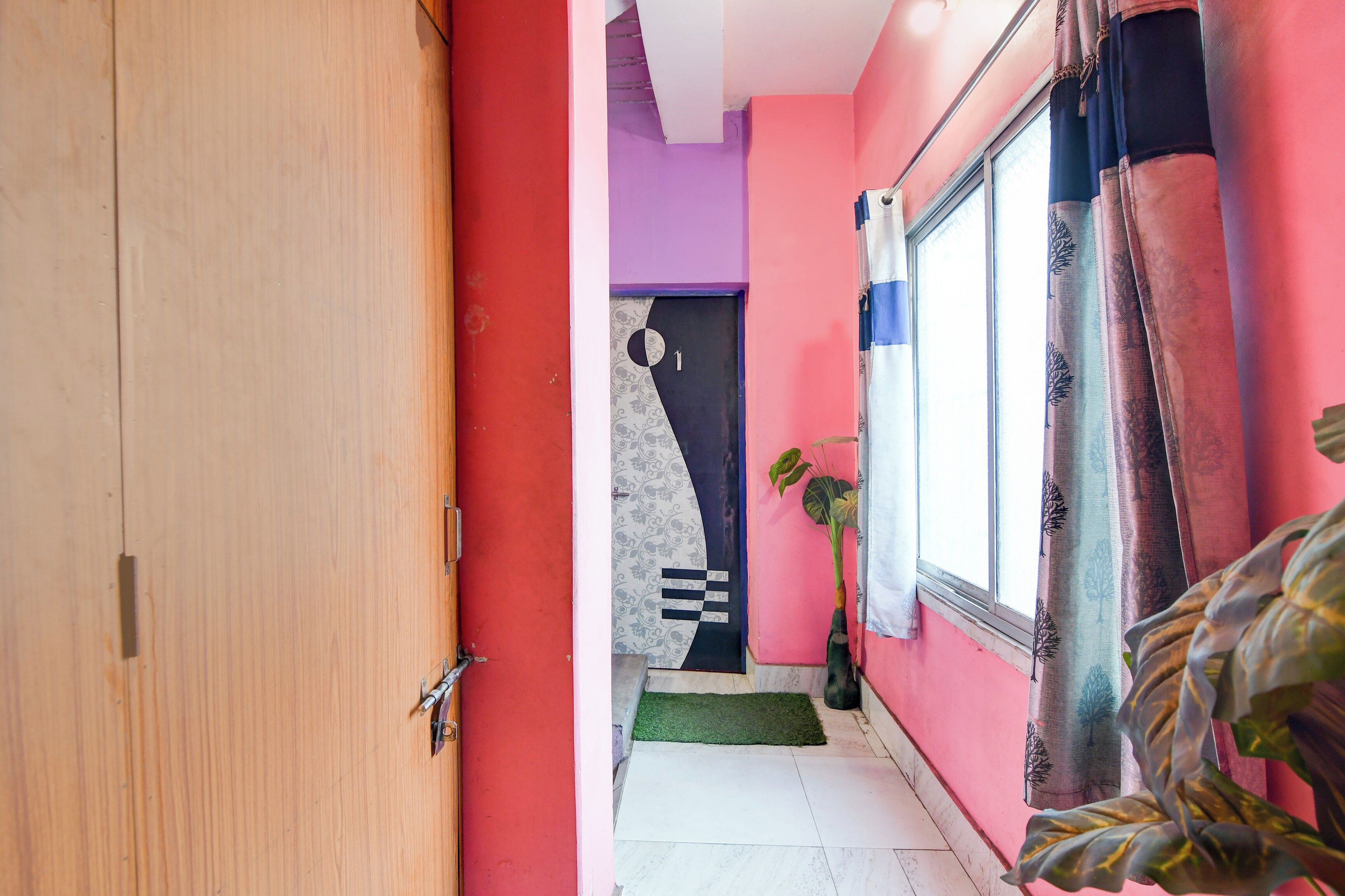 Un couloir étroit avec une porte en bois sur la gauche, un mur violet et une fenêtre avec des rideaux sur la droite.