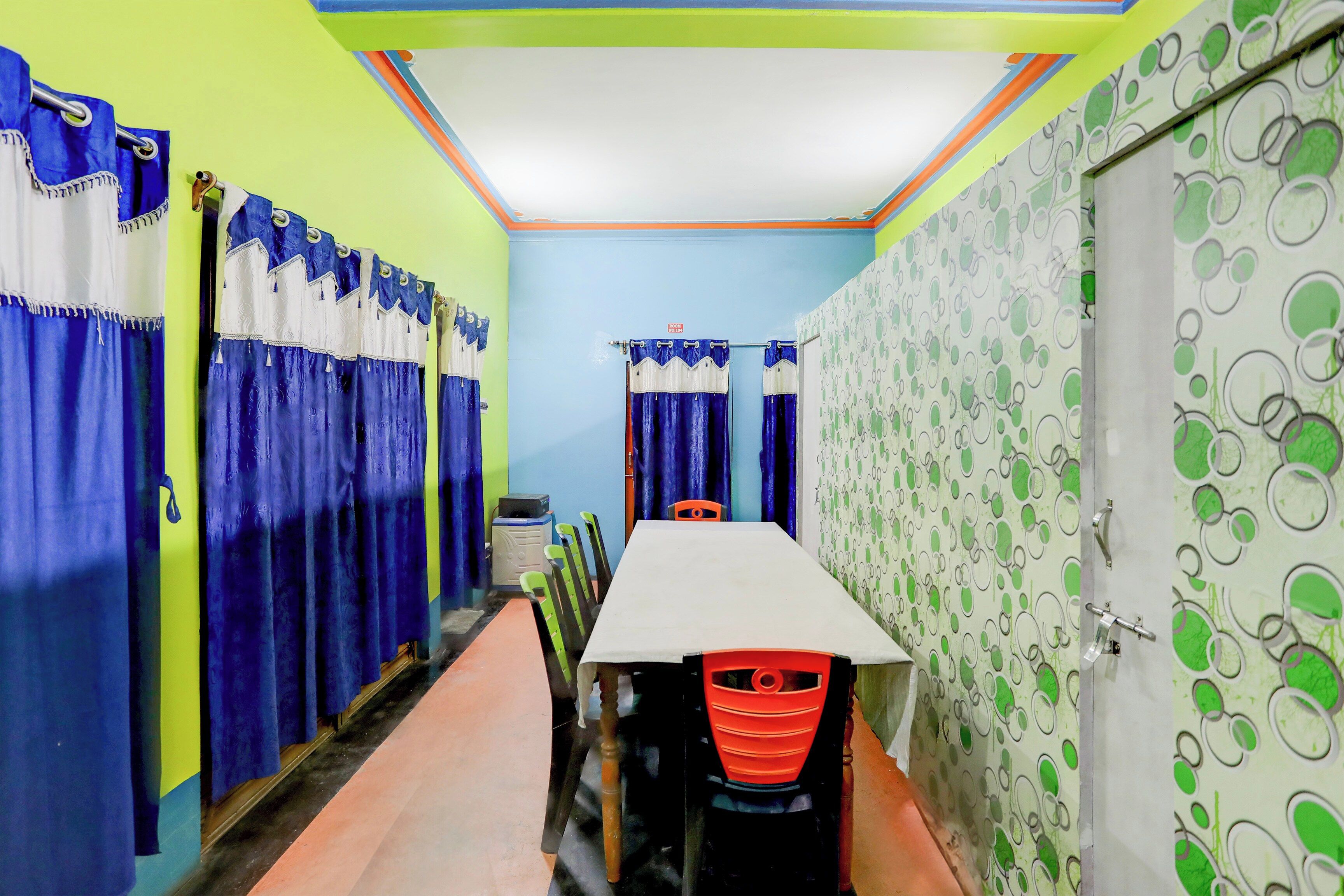 A hotel corridor with a row of rooms, a table with chairs, and a patterned wall.