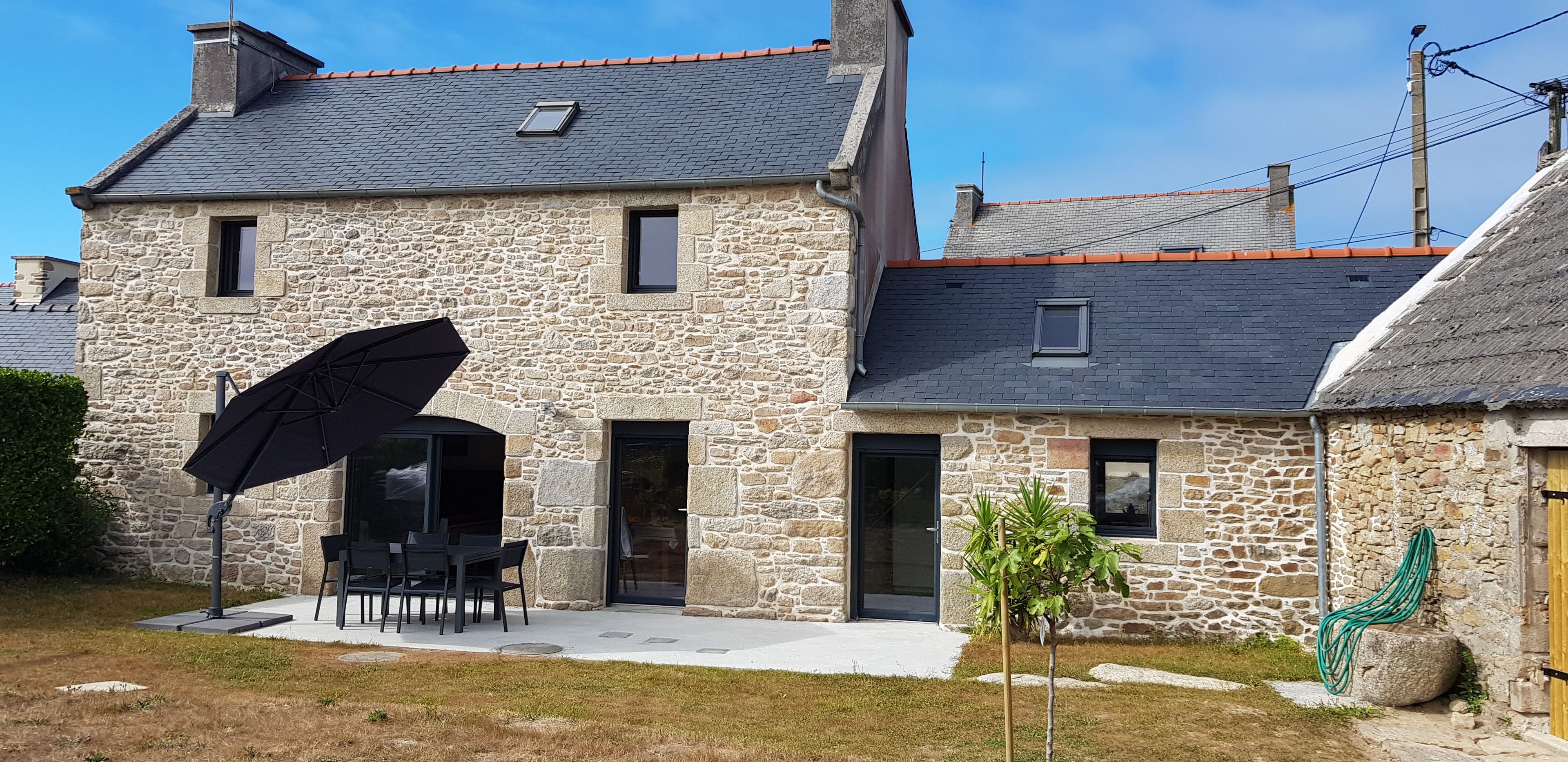 Une maison en pierre avec un patio, un parapluie noir et un petit arbre.