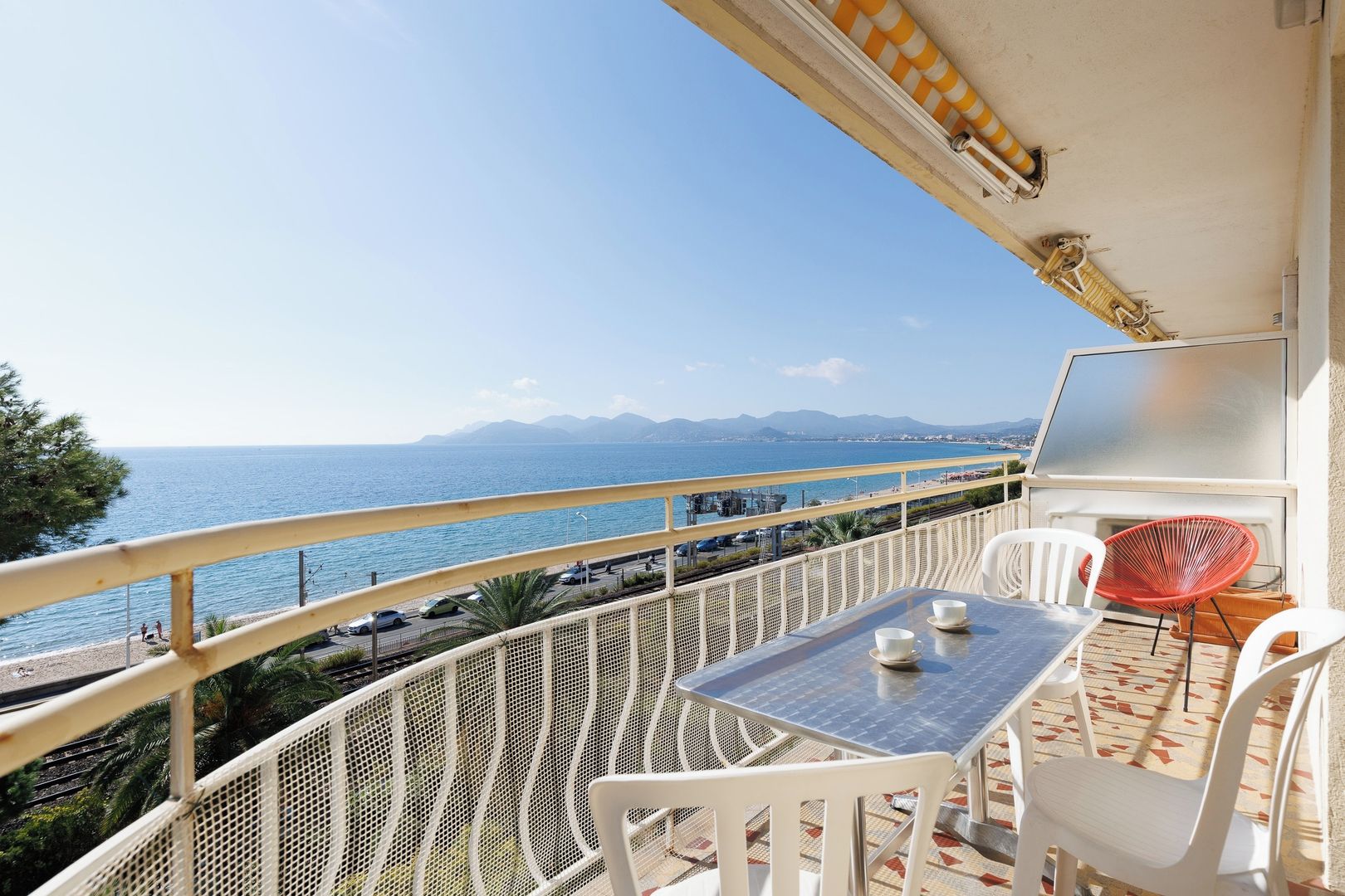 A balcony with a table and chairs, overlooking a coastal view with mountains in the distance.