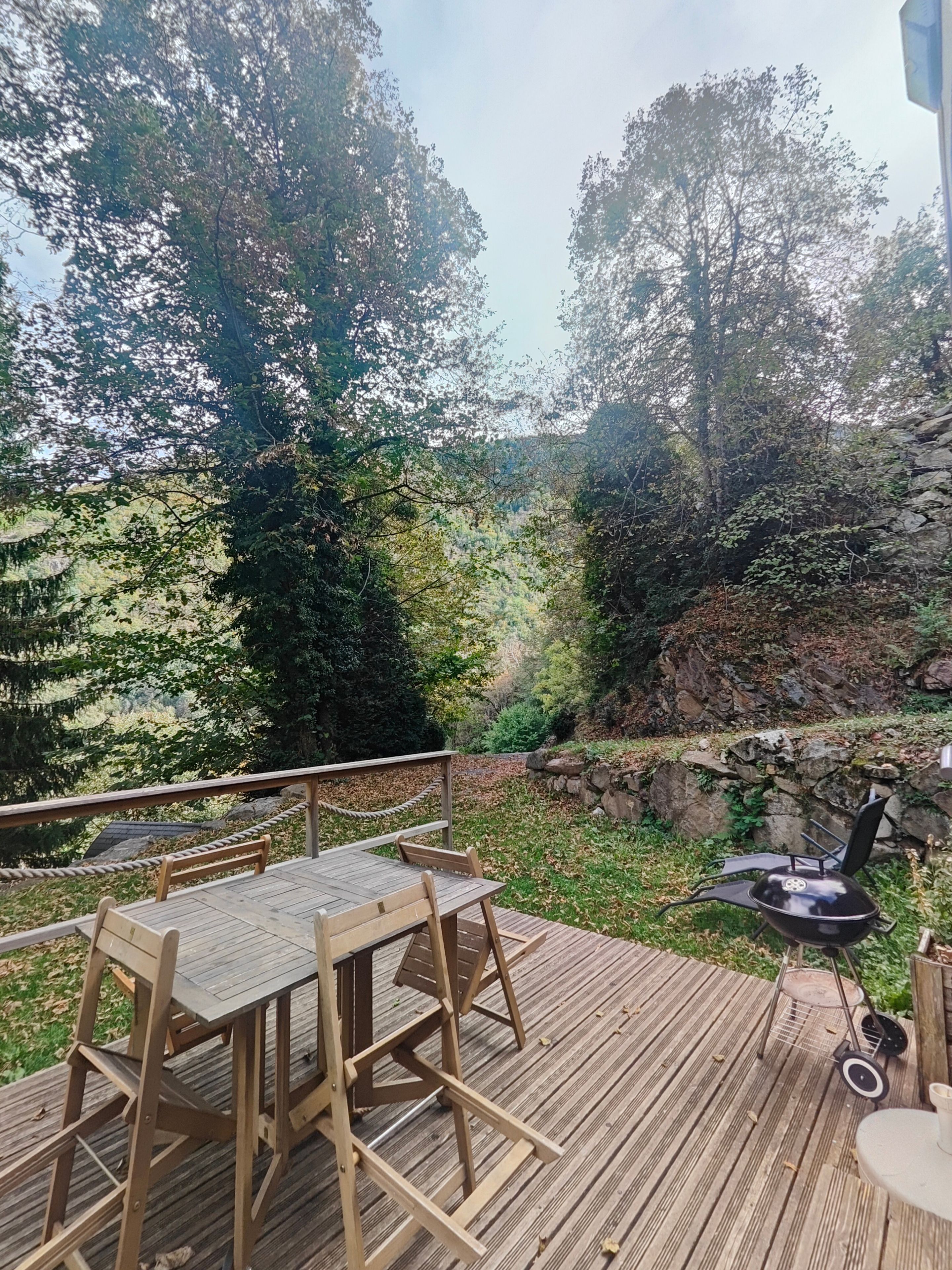 Una terraza al aire libre con una mesa y sillas de madera, una parrilla de barbacoa y vistas a una zona boscosa con un muro de piedra.