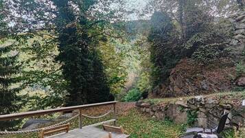 Una terraza al aire libre con una mesa y sillas de madera, una parrilla de barbacoa y vistas a una zona boscosa con un muro de piedra.