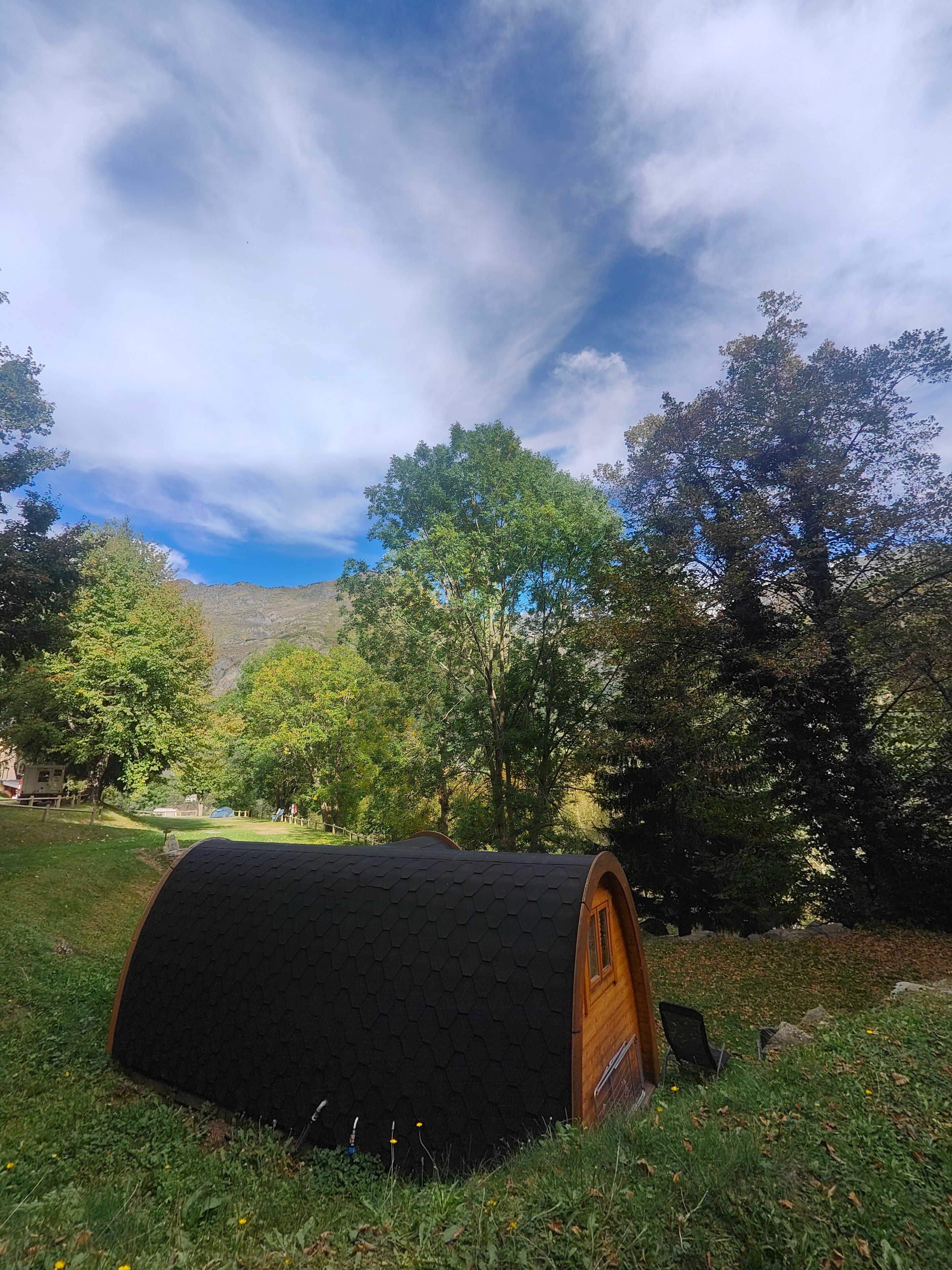 A cylindrical structure with a wooden door, surrounded by grass and trees, with a mountainous backdrop.