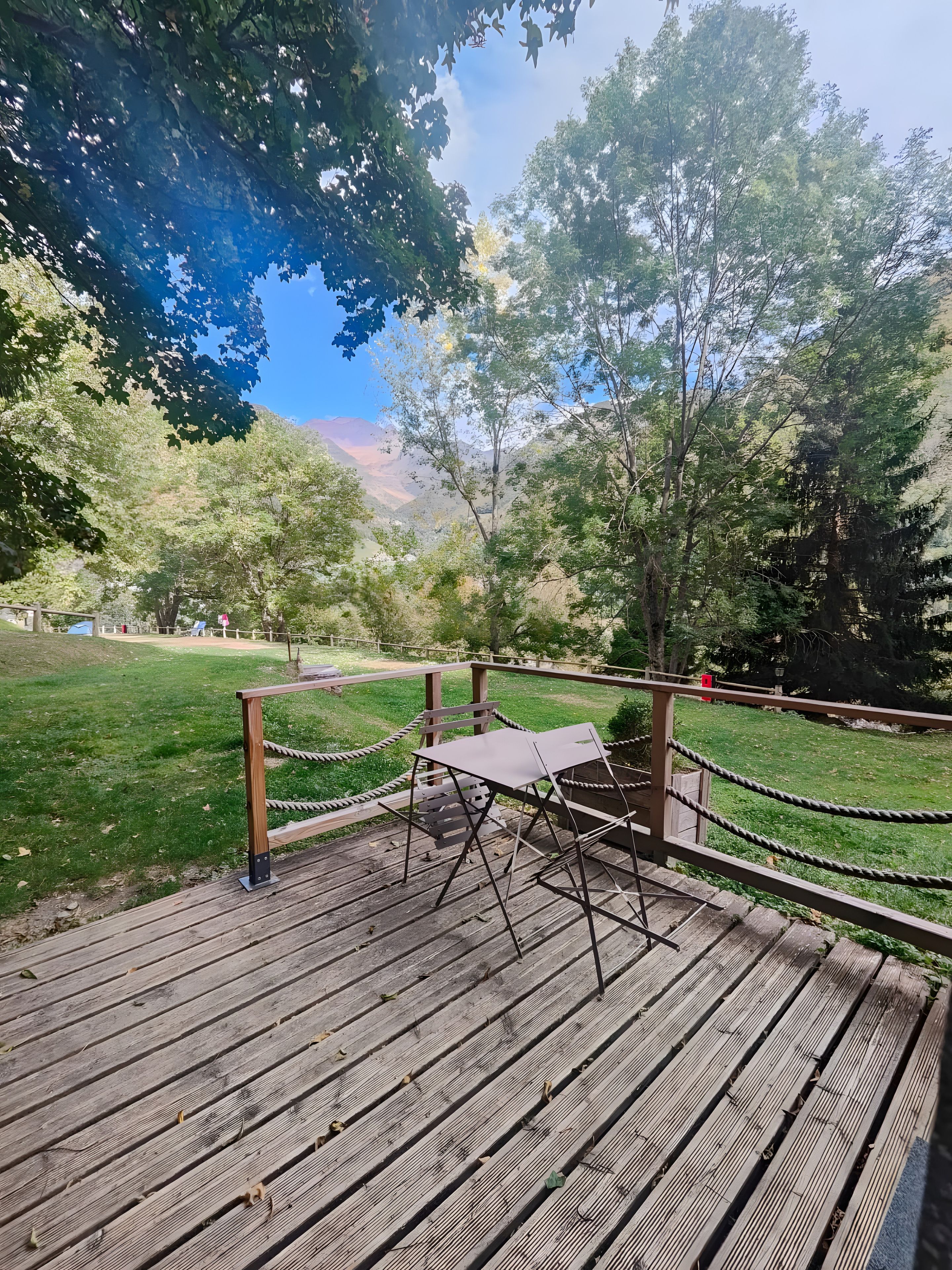 Una terraza de madera con una mesa y sillas de metal, rodeada por una barandilla de cuerda, con vistas a un área verde con árboles y una montaña al fondo.