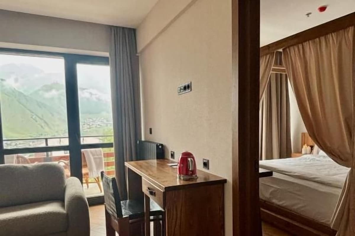 A hotel room with a bed, a desk with a kettle, a sofa, and a view of the outdoors.