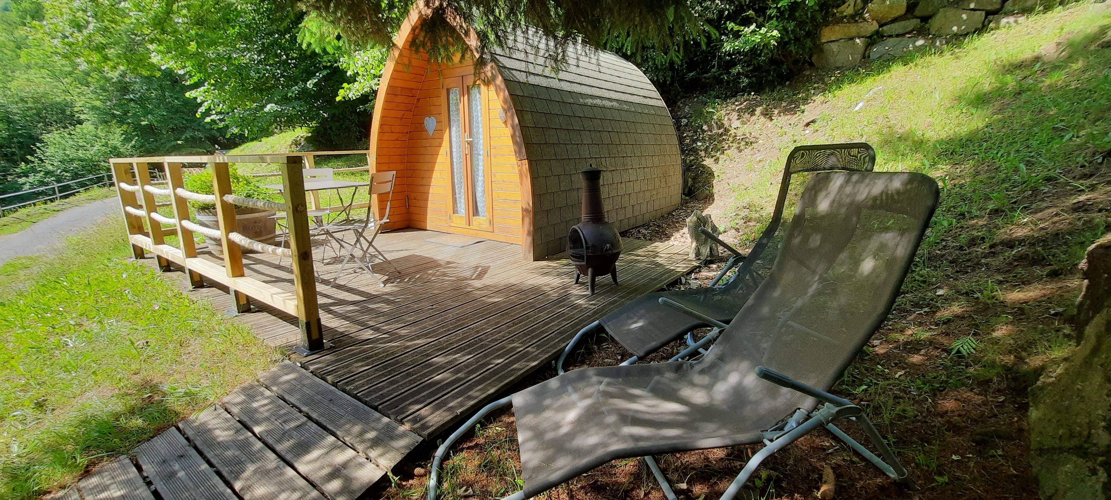 A wooden deck leading to a small cabin with a heart-shaped window, a metal chair, and a stone wall in the background.