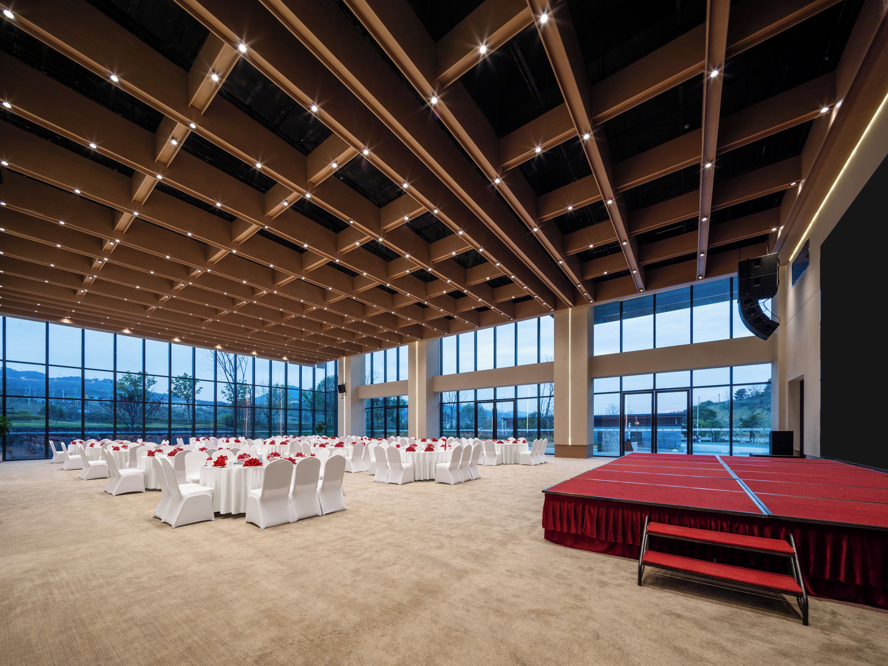 Un salón amplio con escenario, filas de sillas blancas y ventanas grandes.