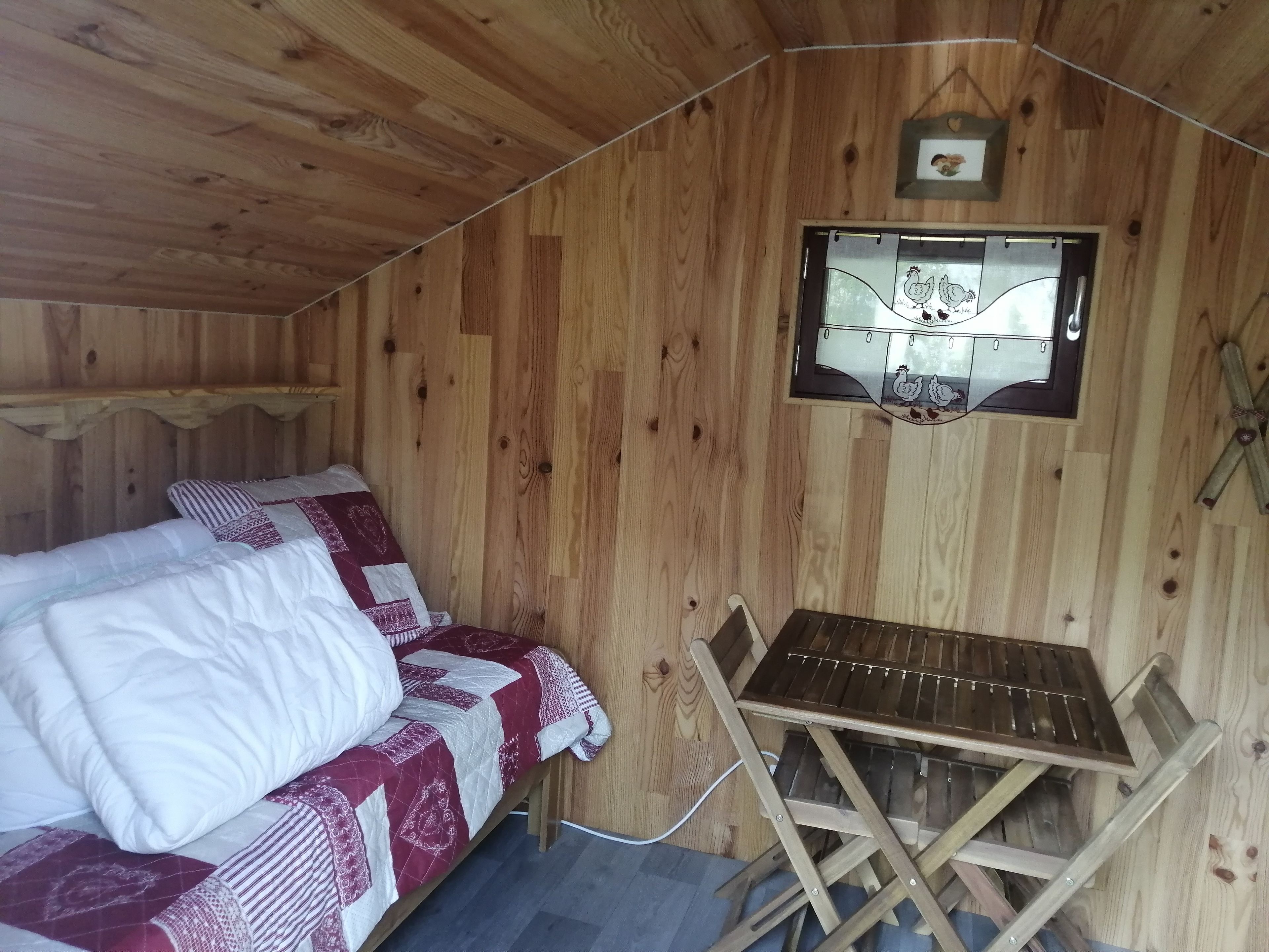 A wooden cabin room with a bed, a small table, and a window with a picture.