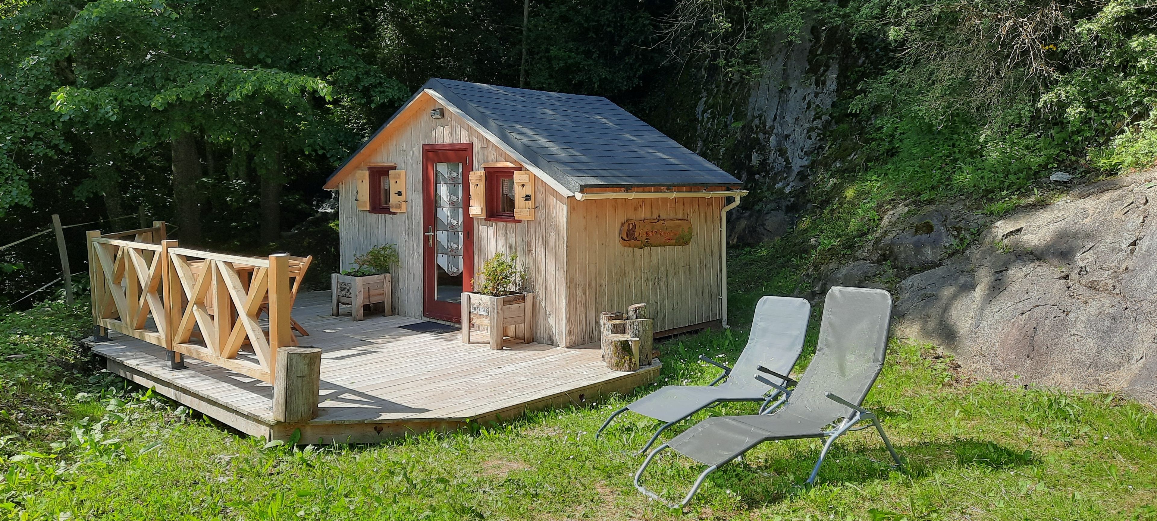 Una cabaña de madera con puerta roja, un porche pequeño y una terraza con sillas.