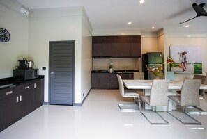 A modern kitchen with dark cabinets, a dining area with a white table and chairs, and a painting on the wall. - Hill Retreat 3BR Pool Villa near Nai Harn, Rawai (Tambon Rawai)