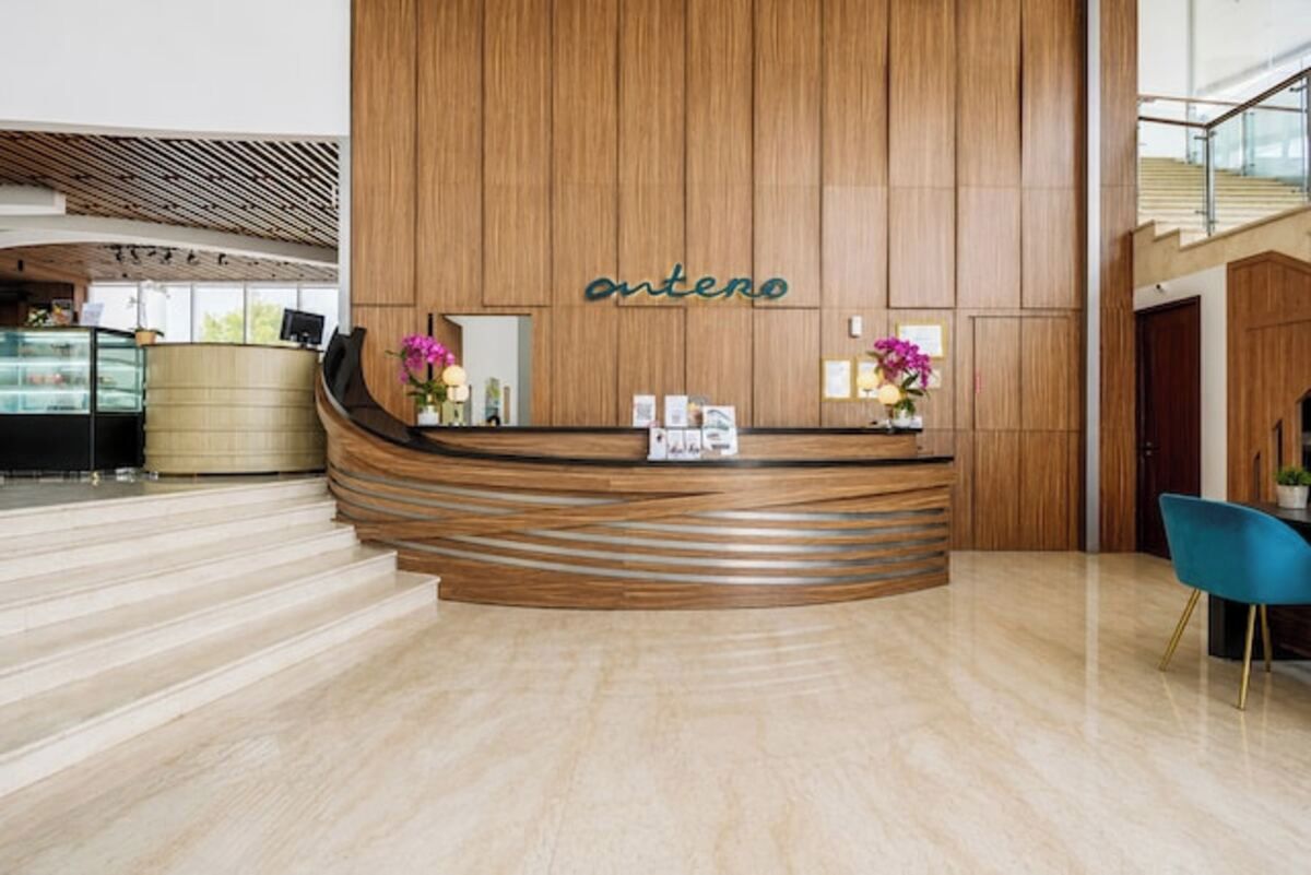Un mostrador de recepción con detalles de madera y la palabra 'antero' en la pared. Hay escalones que conducen al mostrador, una silla azul y una escalera al fondo.