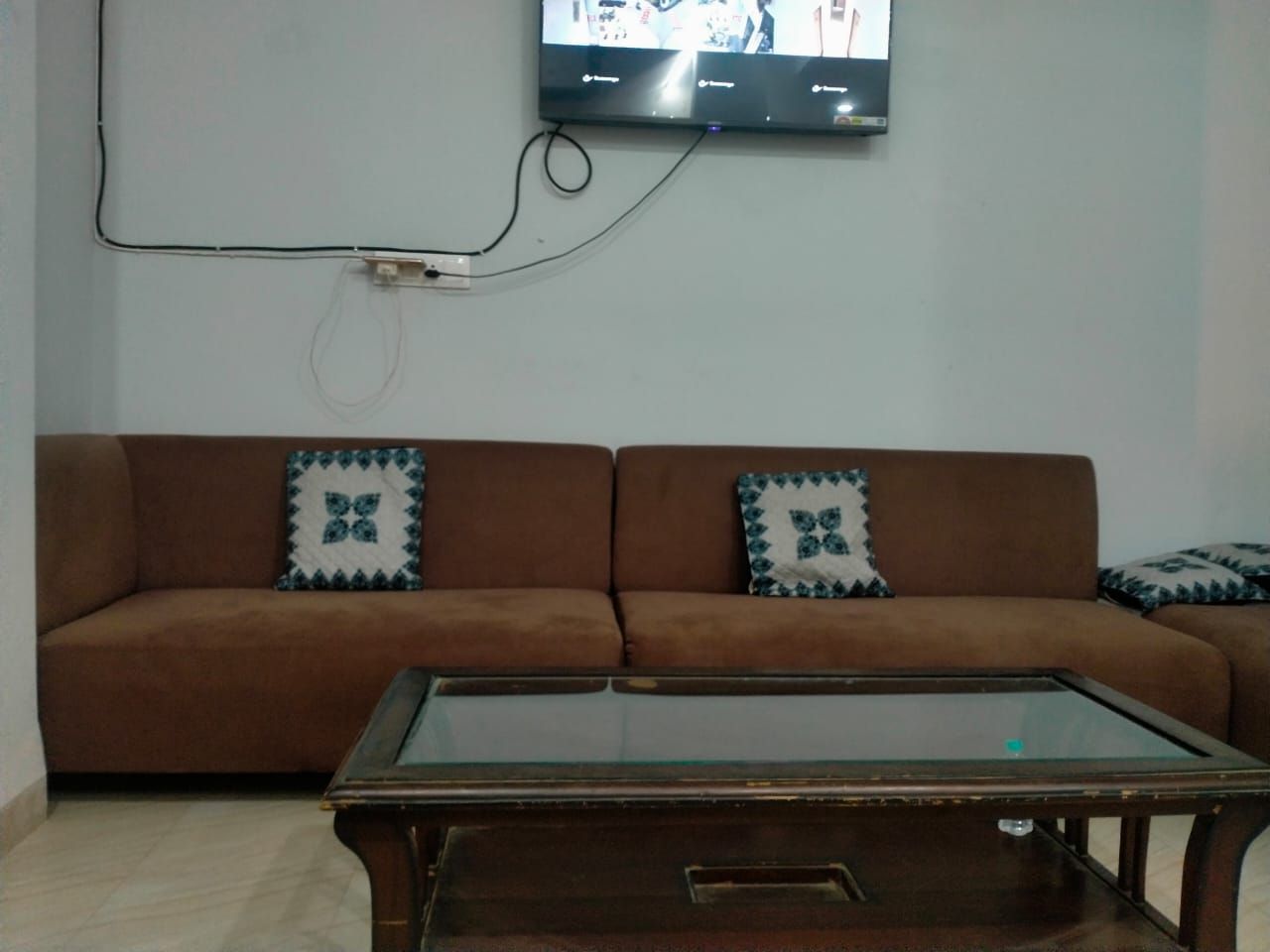 A living room with a brown sofa, two patterned cushions, a glass-top coffee table, and a flat-screen TV mounted on the wall.