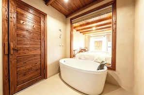 Een badkamer met een houten deur, een witte badkuip en een spiegel die een slaapkamer met een bed en een stoel weerspiegelt.