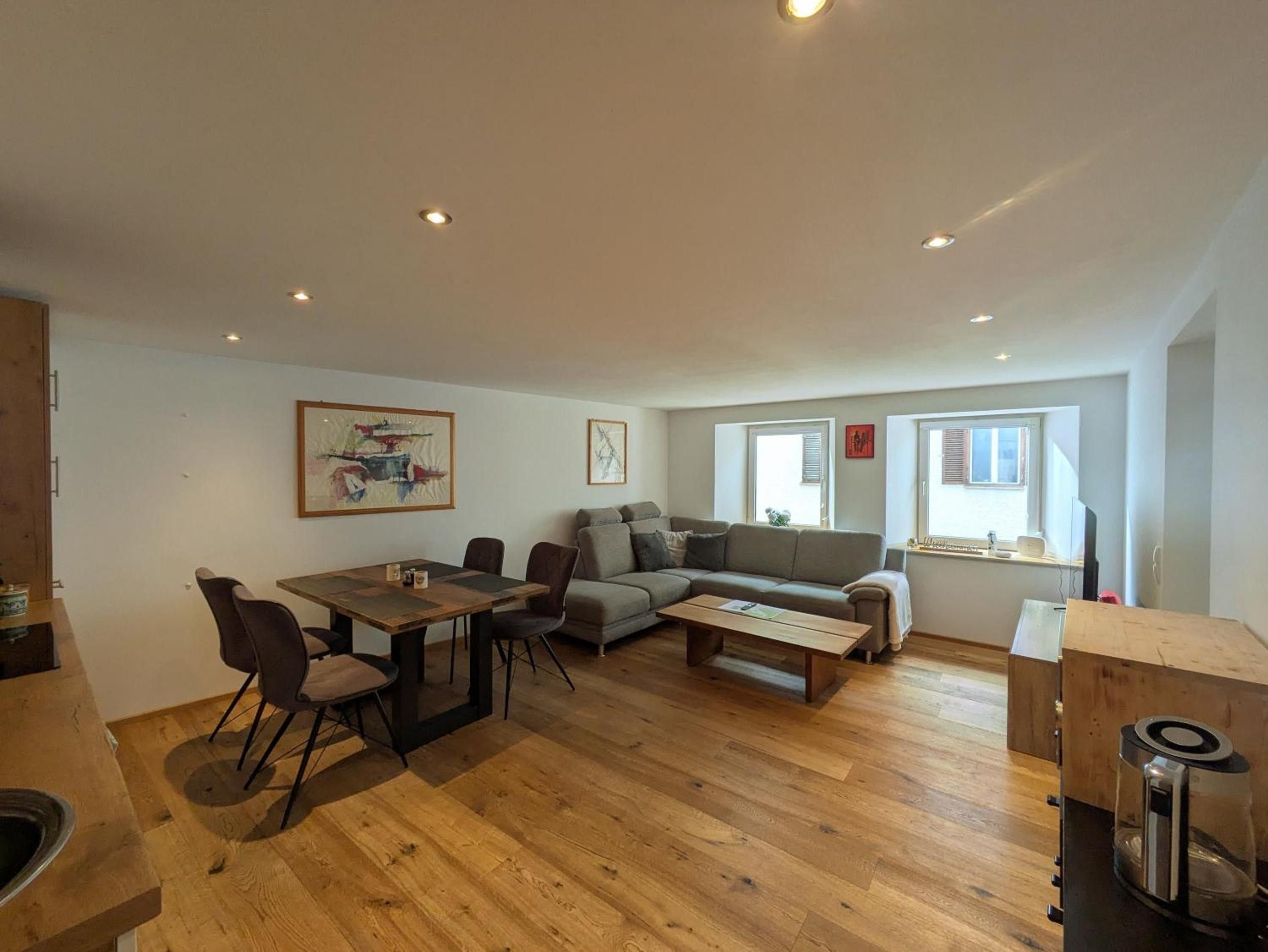 Un salon moderne avec un plancher en bois, une table à manger avec des chaises, un canapé et une table basse. Il y a deux fenêtres avec des stores, un tableau accroché au mur et, au fond, on aperçoit un coin cuisine.