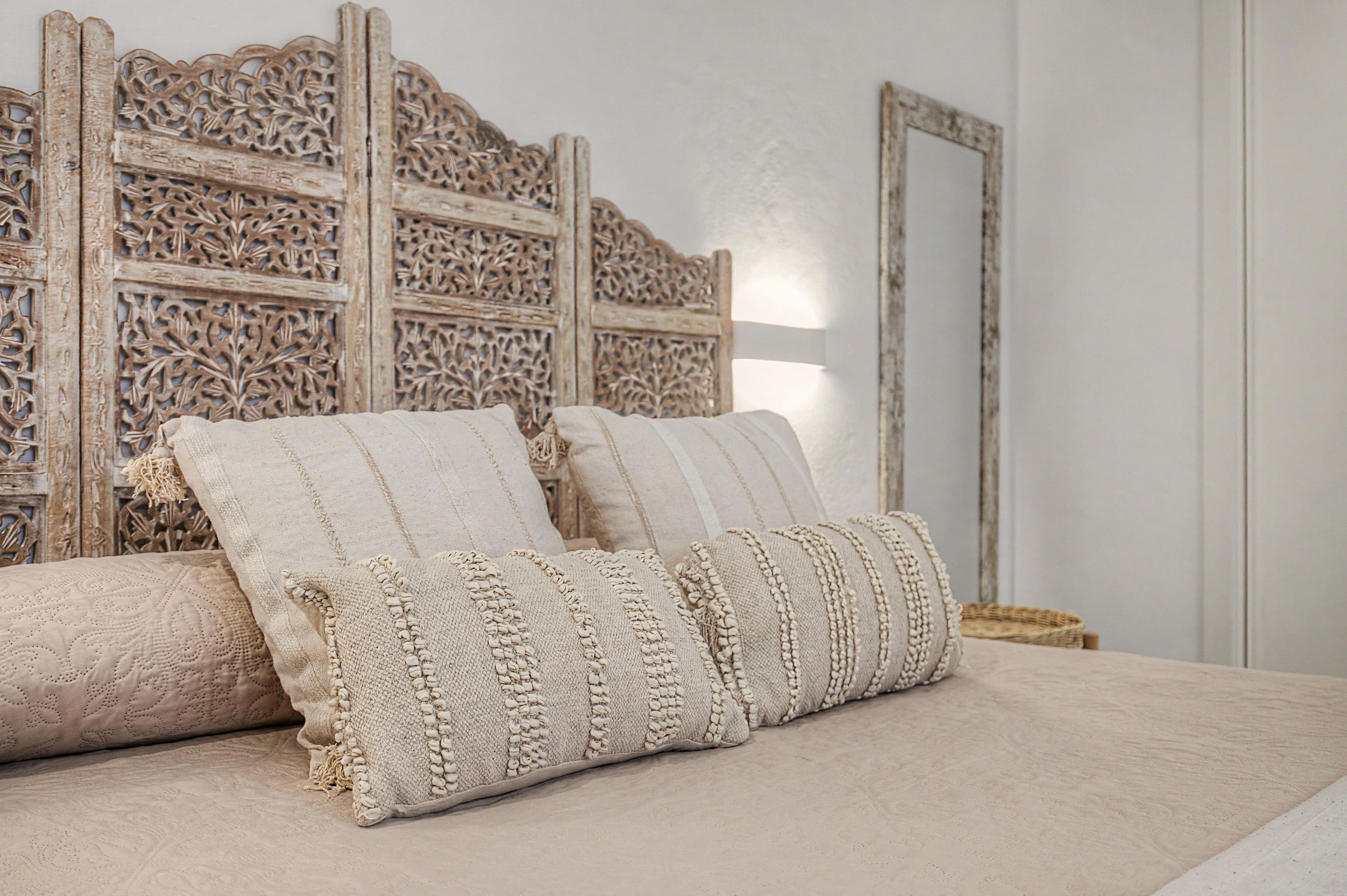 Een bed met een gebobbelde beige hoes en decoratieve kussens, een houten hoofdbord met gedetailleerde houtsnijwerken, en een spiegel aan de muur.
