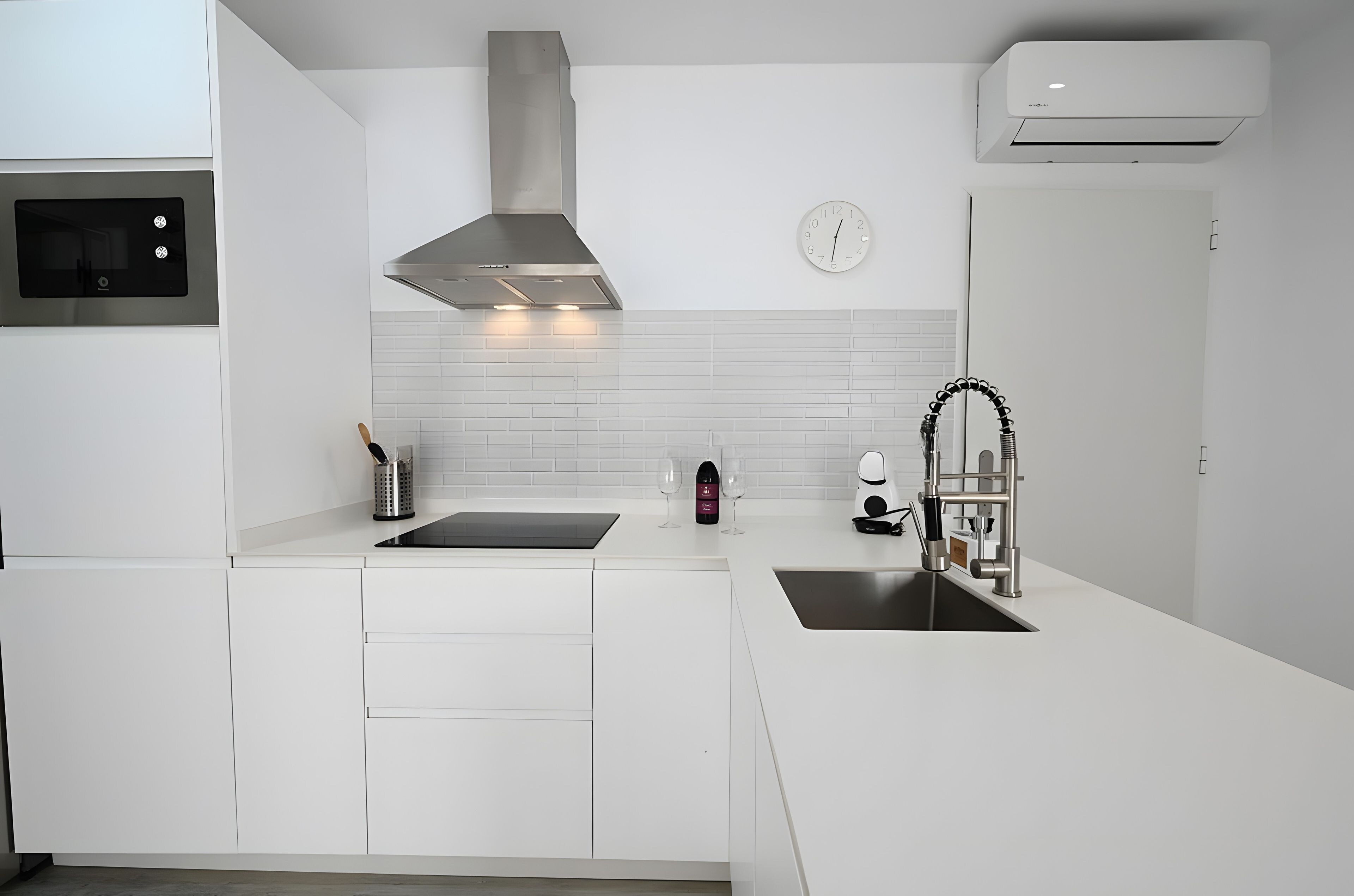 A modern kitchen with white cabinetry, a stainless steel range hood, a built-in oven, a double sink, and a wall-mounted clock.