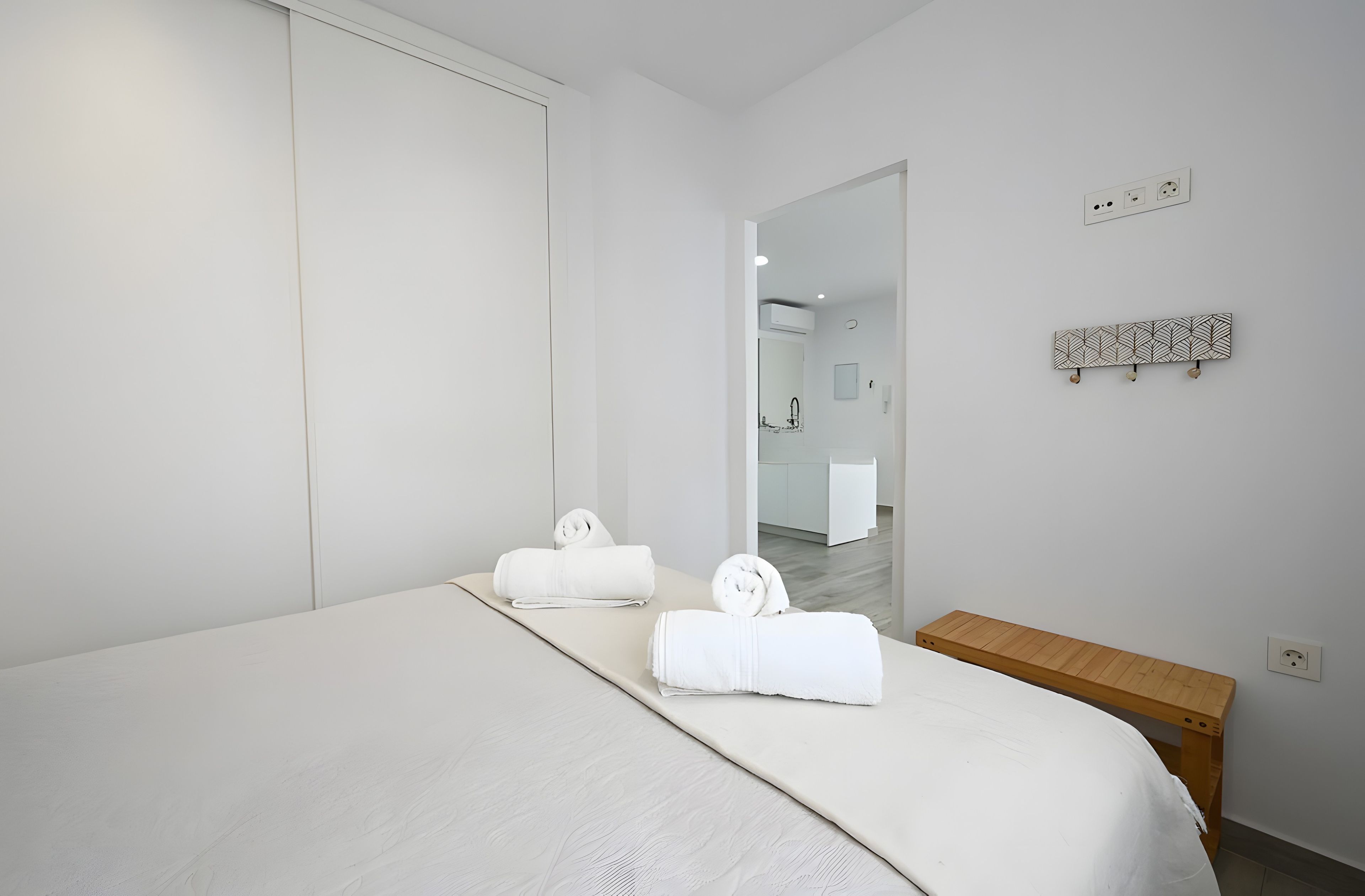 A modern hotel room with a large bed, two towels, a wooden bench, a mirror, and a wall-mounted light fixture.