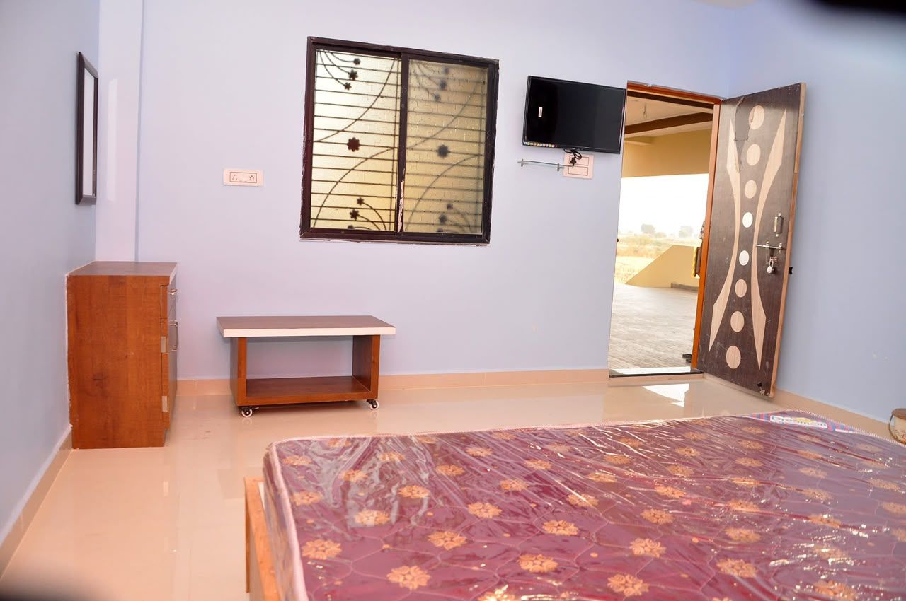 Une chambre à coucher comprenant un lit, une armoire en bois, une petite table, un téléviseur fixé au mur et une porte donnant sur une autre pièce.