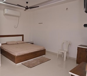 Quarto com cama de madeira, uma cadeira branca, uma mesinha e um ventilador de teto.