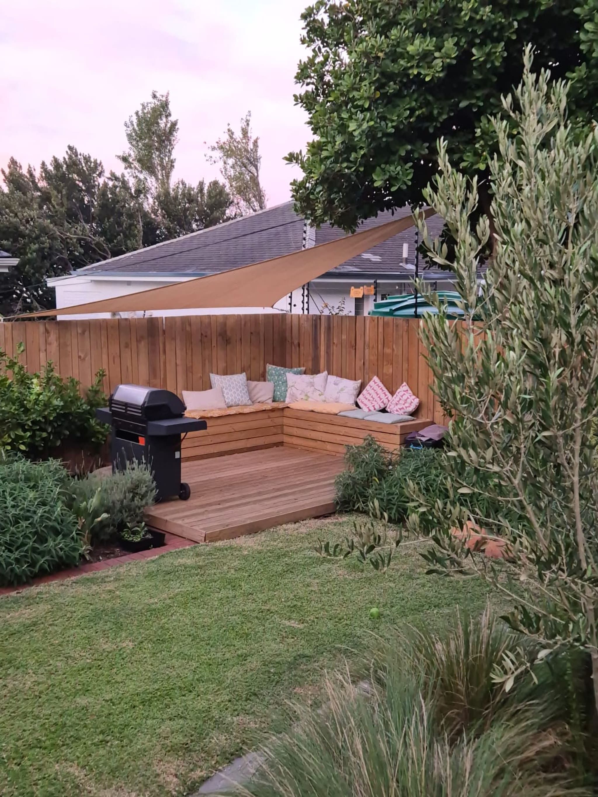 Een achtertuin met een houten terras, een barbecue en een zithoek met kussens.