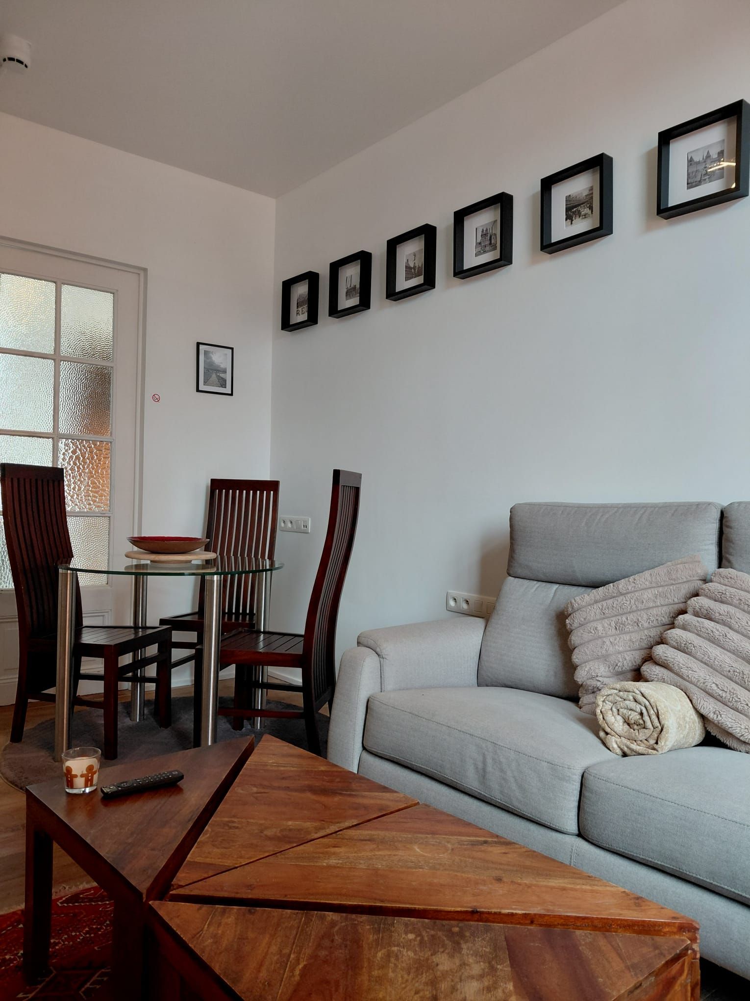 A modern living room with a glass dining table, two wooden chairs, a grey sofa with a throw blanket, and a wooden coffee table.