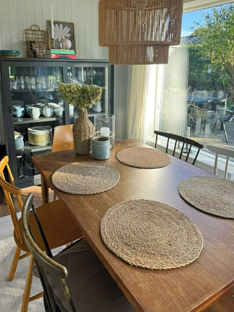 Une table à manger agrémentée de sets de table en fibres tressées, un vase avec des plantes vertes et un luminaire suspendu.