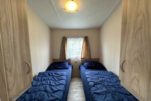 A narrow room with two beds, wooden wardrobes, a window with curtains, and a ceiling light.