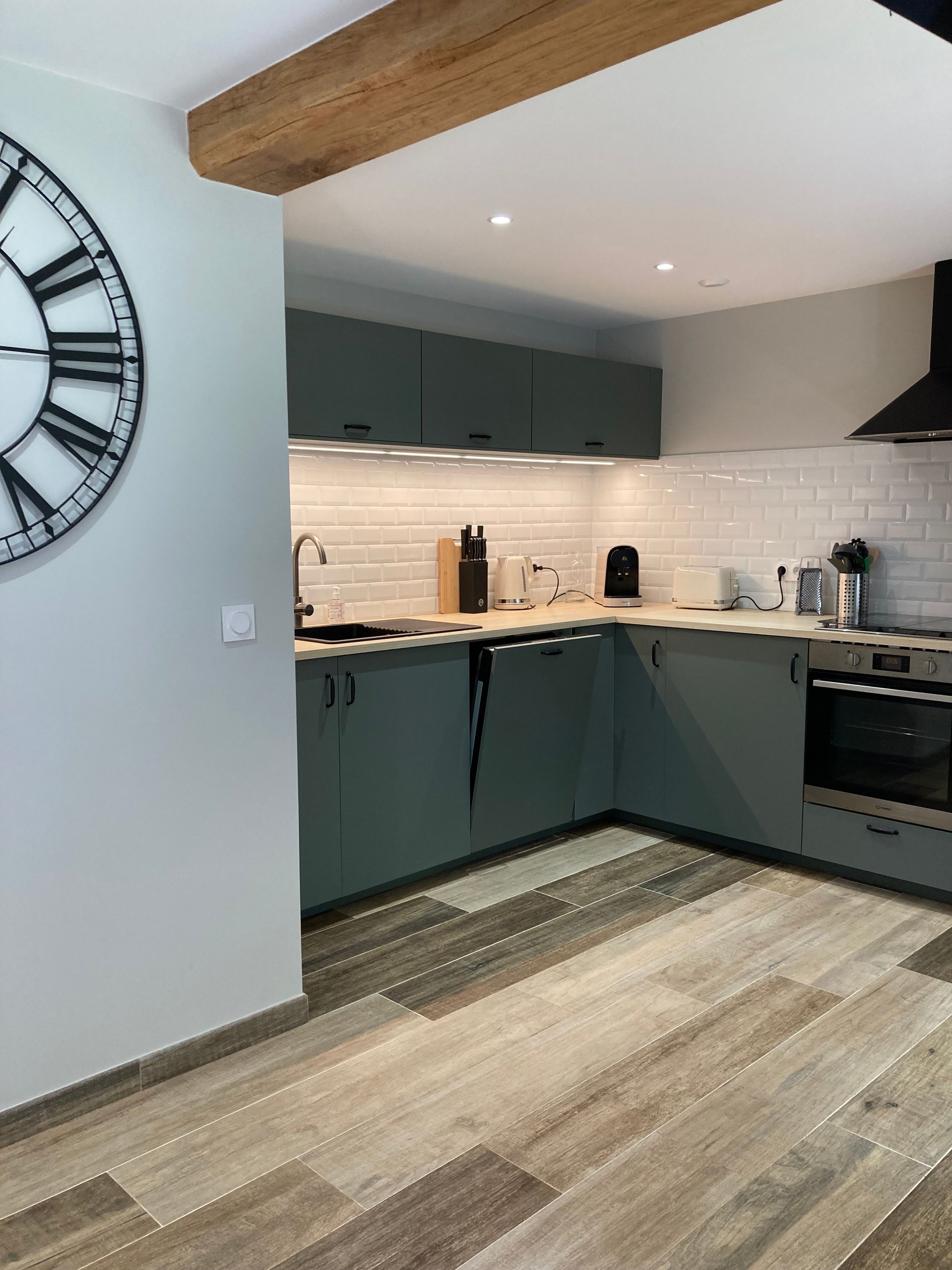 A modern kitchen with a large clock, grey cabinets, a wooden ceiling, and a tiled backsplash.
