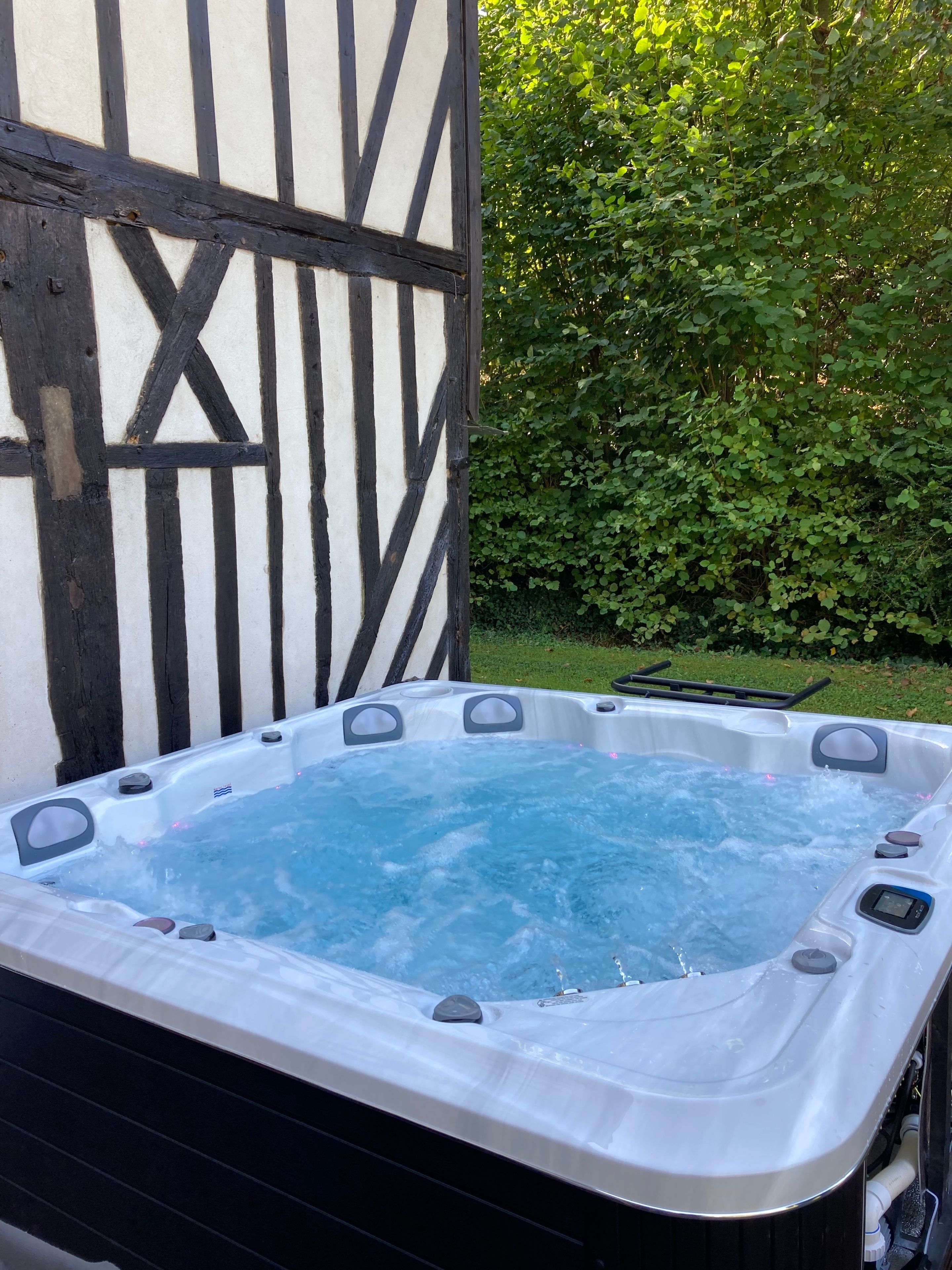 A hot tub with a control panel, set against a backdrop of a half-timbered building and a green hedge.
