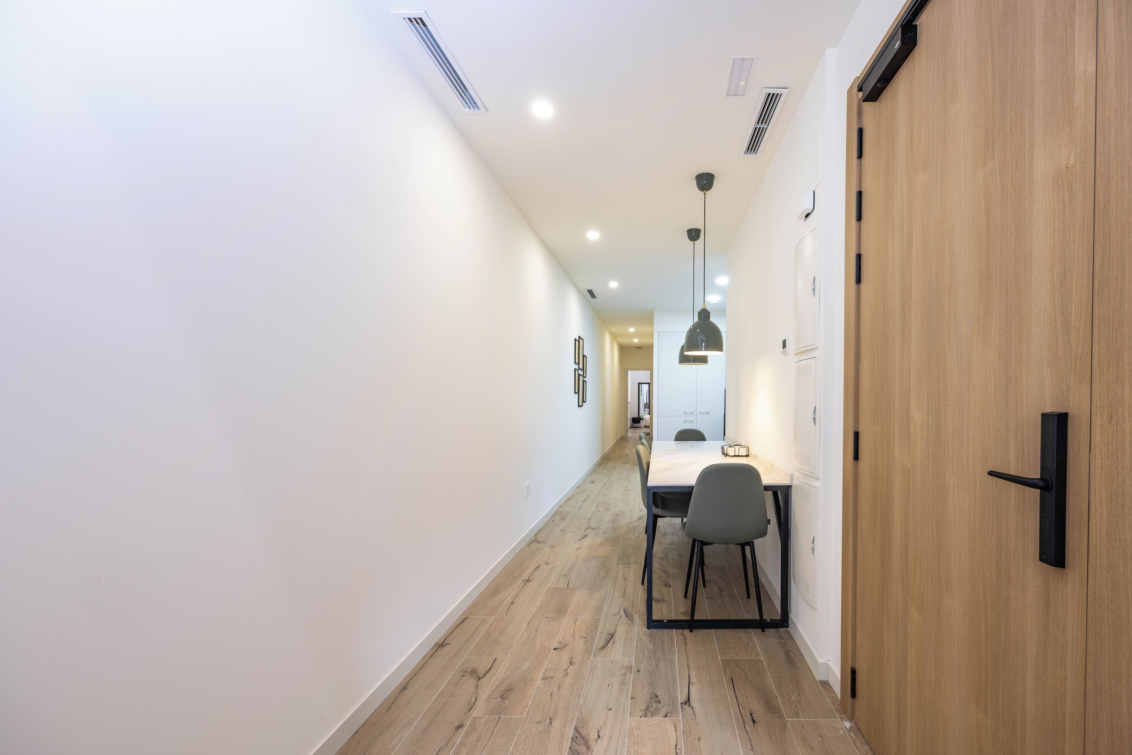 Un couloir avec une porte en bois, une table à manger avec des chaises et des luminaires encastrés au plafond.