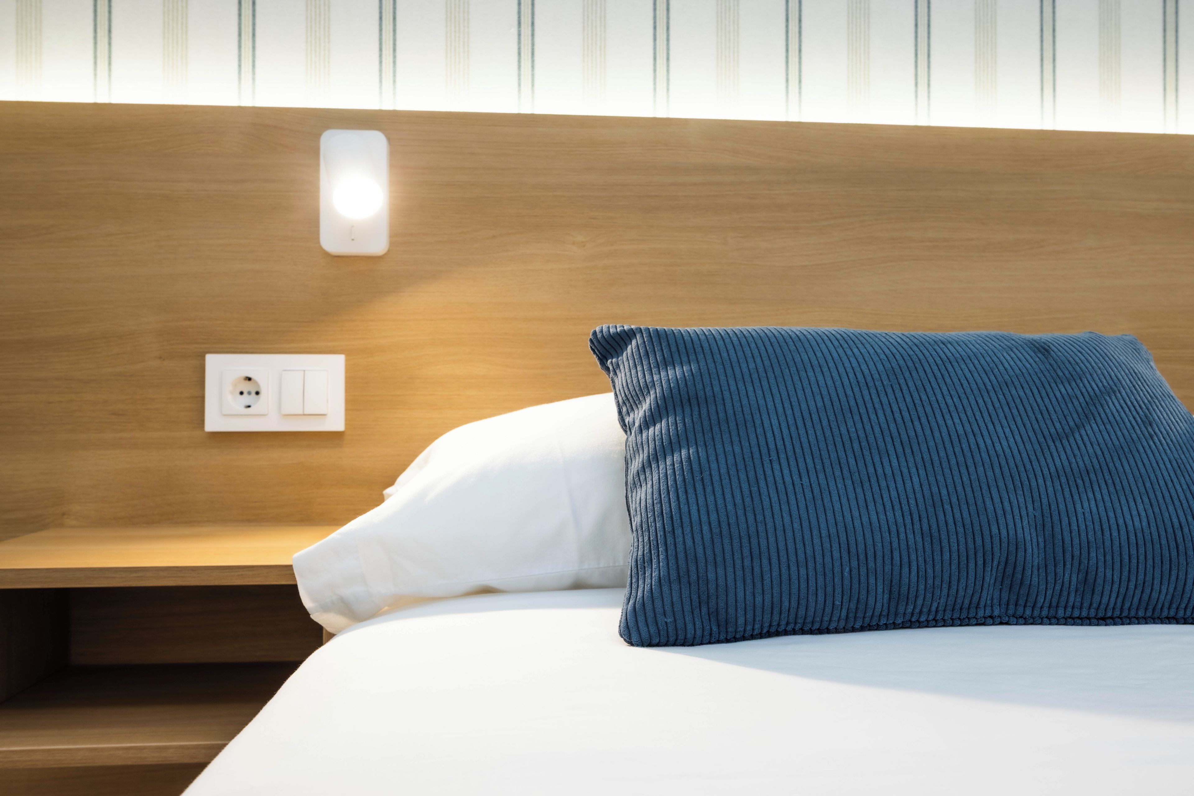 A neatly made bed with a white pillow and a blue pillow, a wooden headboard, and a wall socket.