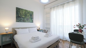 A neatly made bed with white linens and towels, a bedside lamp, a small table with a lamp, a chair, a desk with a vase of flowers, and a painting on the wall. - Italianway Easy - Porta Nuova 52 D (Milan)