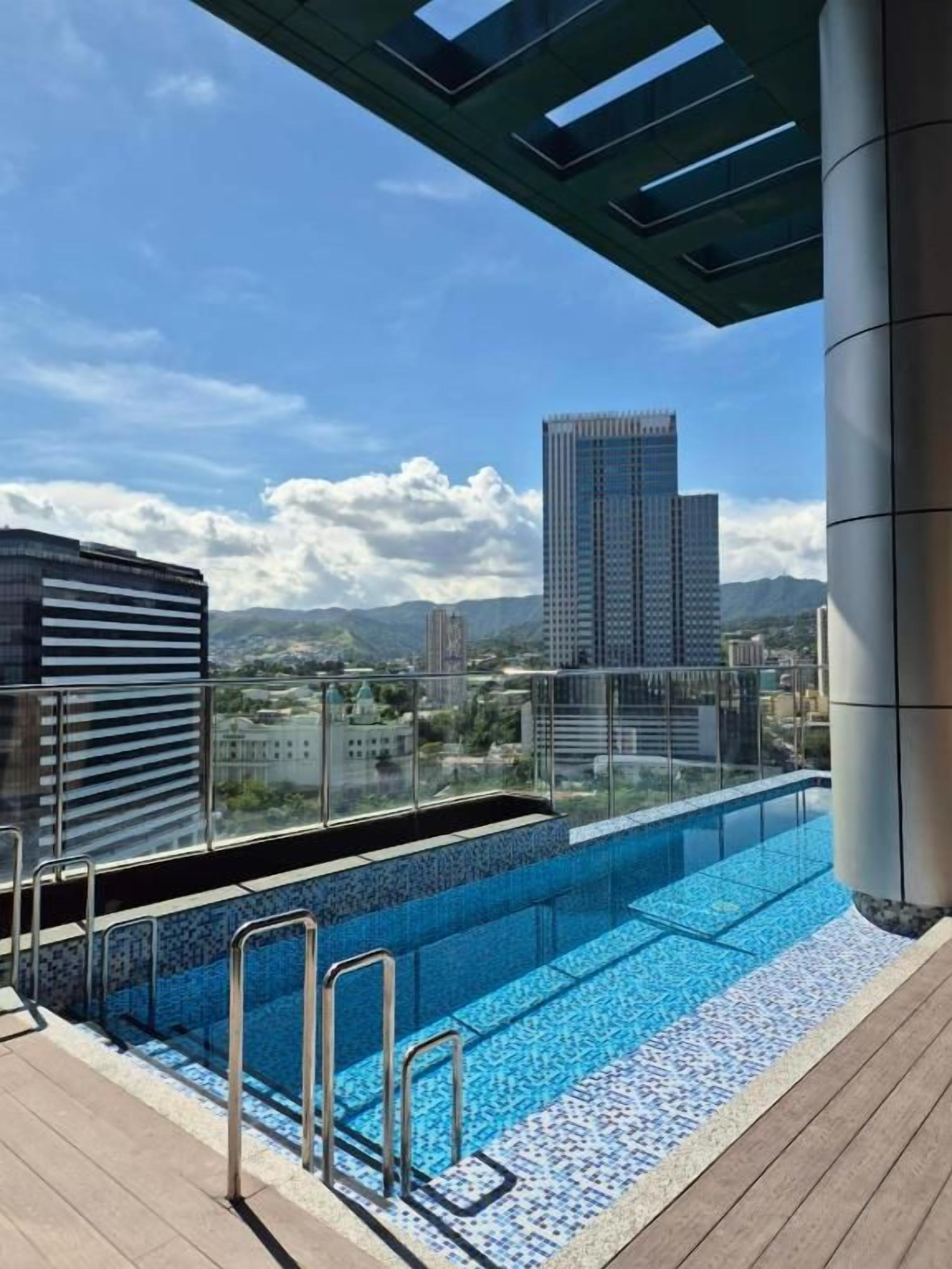 Una piscina en la azotea con vista a la ciudad, rodeada de edificios y un cielo despejado.