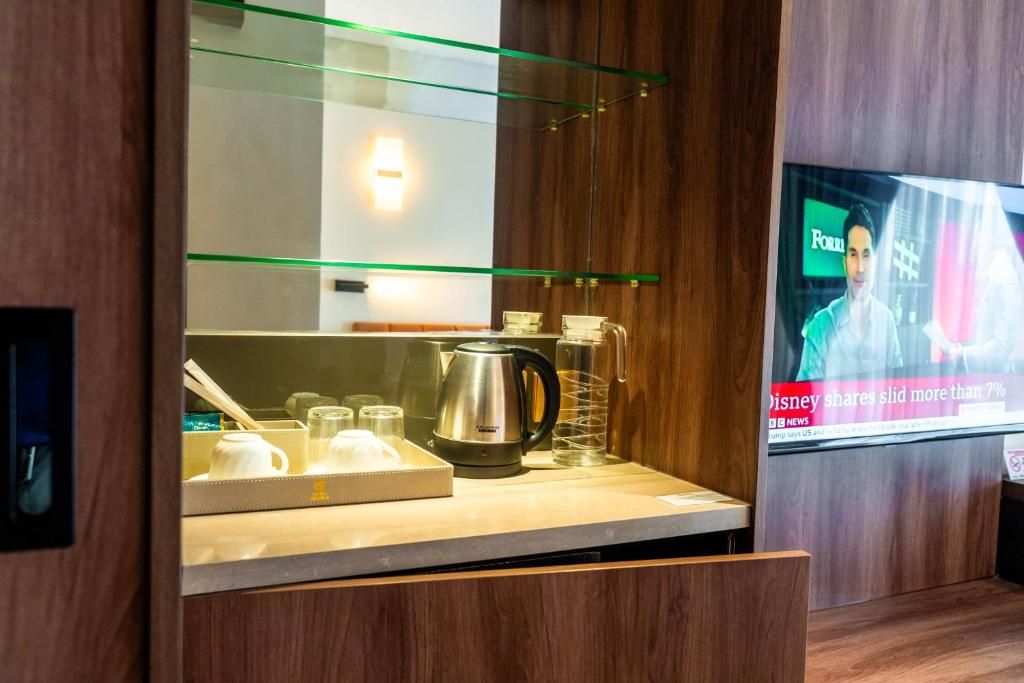 A modern hotel room with a kitchenette area, a television displaying news, and a wooden cabinet.