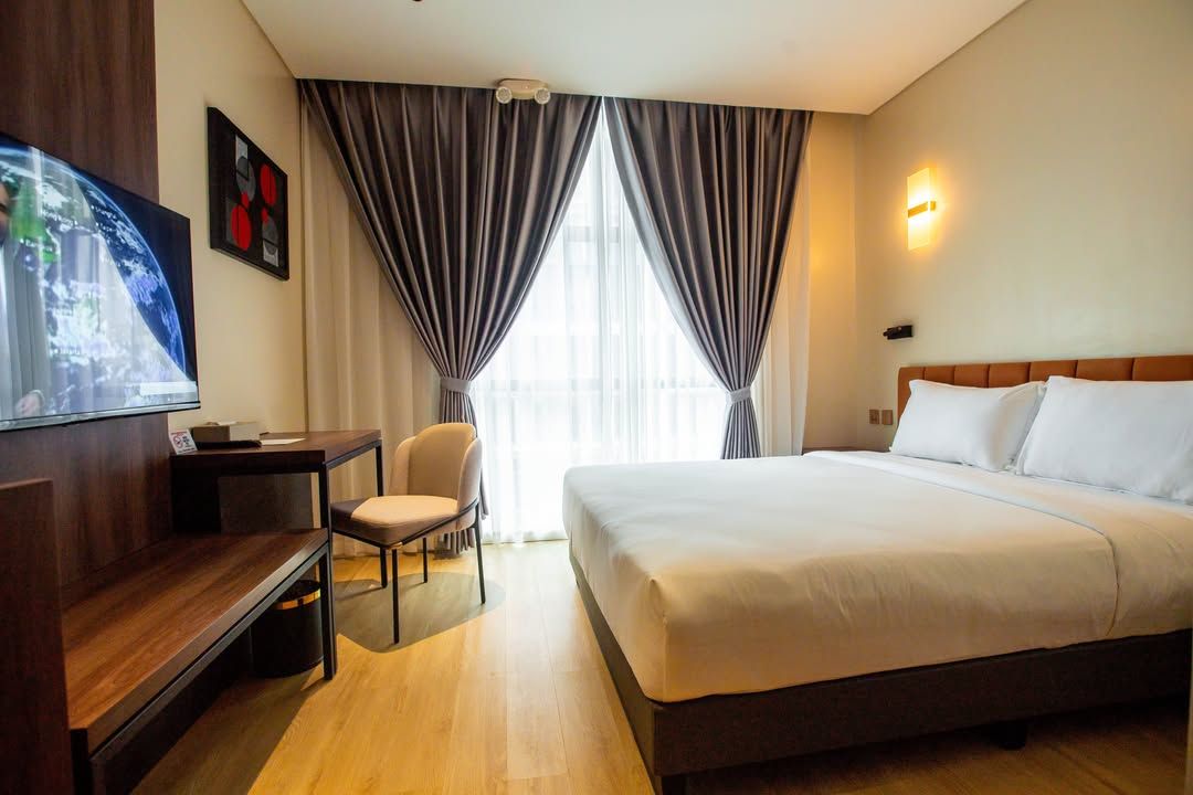 Quarto de hotel com cama, escrivaninha, cadeira, televisão e cortinas.