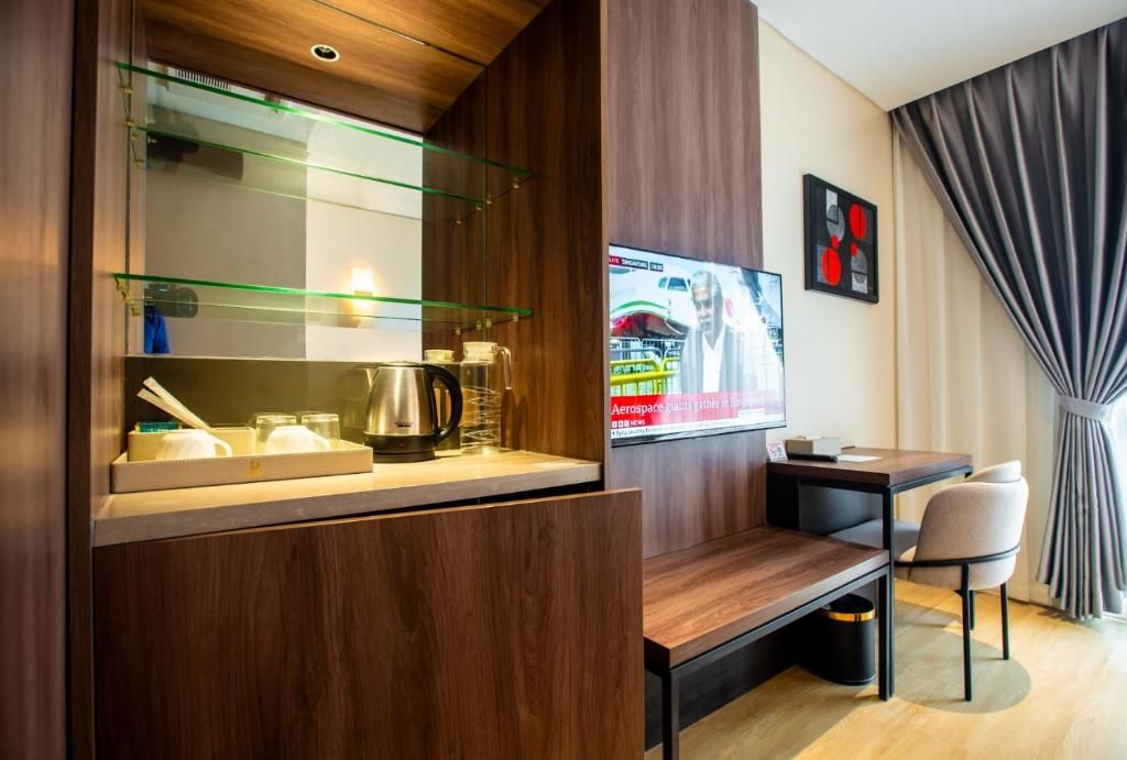 A modern hotel room with a wooden cabinet, a glass shelf with kitchenware, a TV mounted on the wall, and a desk with a chair.