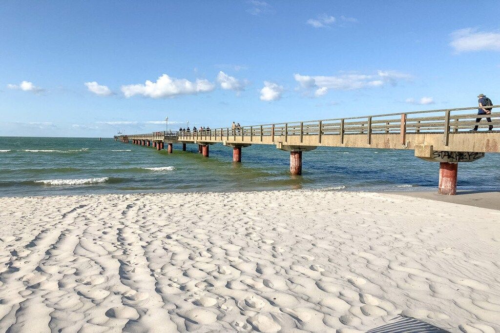 Una playa arenosa con un muelle que se adentra en el agua, con un paseo y una persona caminando.
