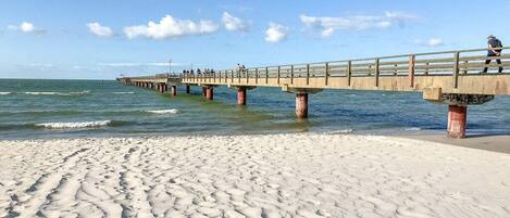 Una playa arenosa con un muelle que se adentra en el agua, con un paseo y una persona caminando.