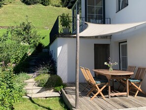 A wooden deck with a table and chairs, surrounded by greenery and a white building. - New Listing (Moos)