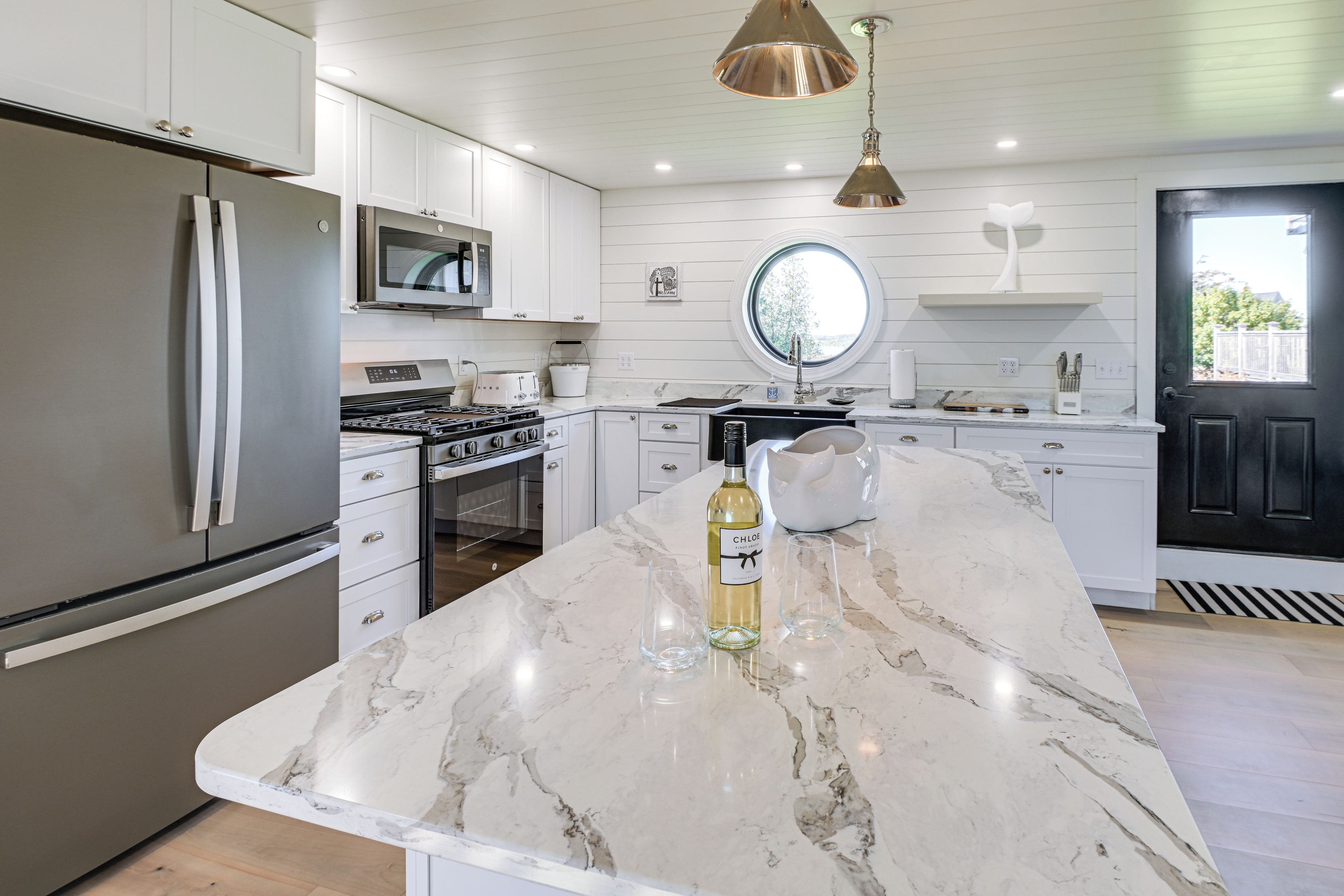 A modern kitchen with white cabinetry, stainless steel appliances, a marble countertop, and a round window above the sink.