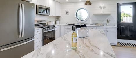 A modern kitchen with white cabinetry, stainless steel appliances, a marble countertop, and a round window above the sink.