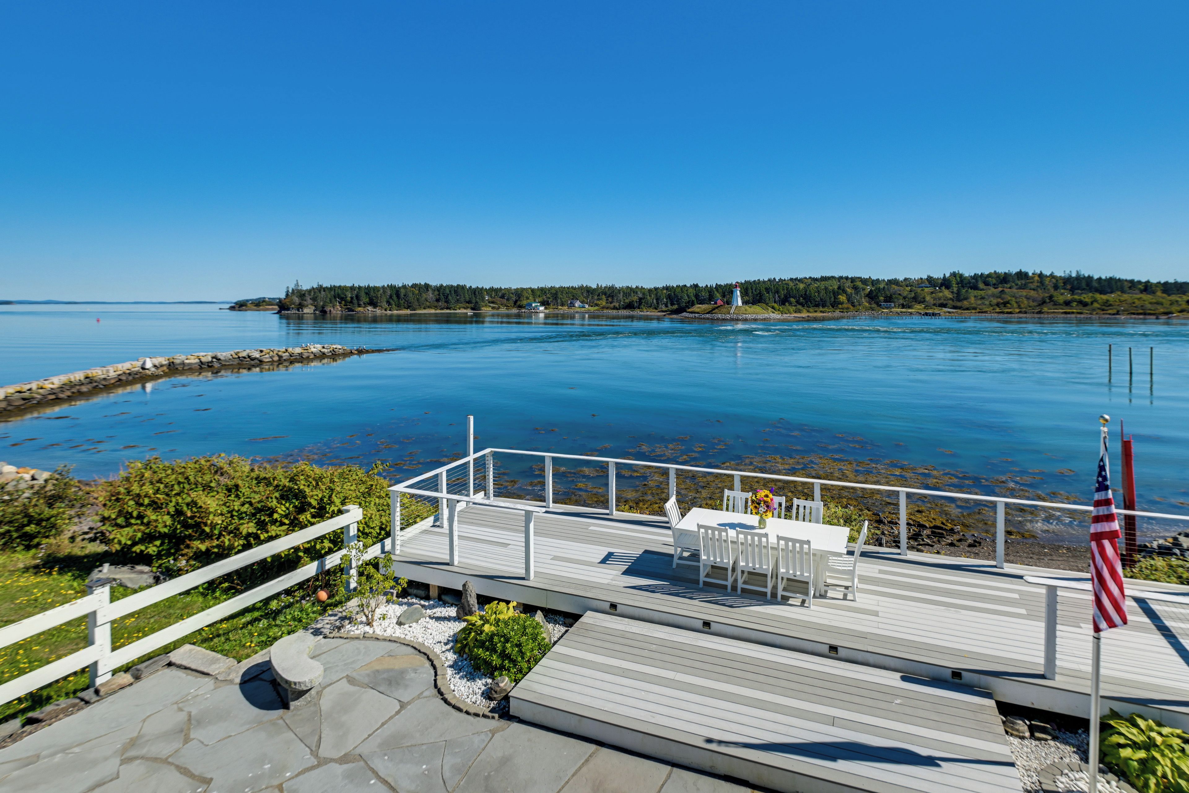 A deck with a table and chairs, an American flag, and a view of a calm body of water with a distant shoreline.