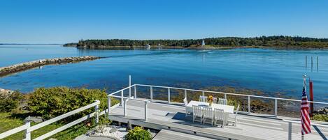 A deck with a table and chairs, an American flag, and a view of a calm body of water with a distant shoreline.